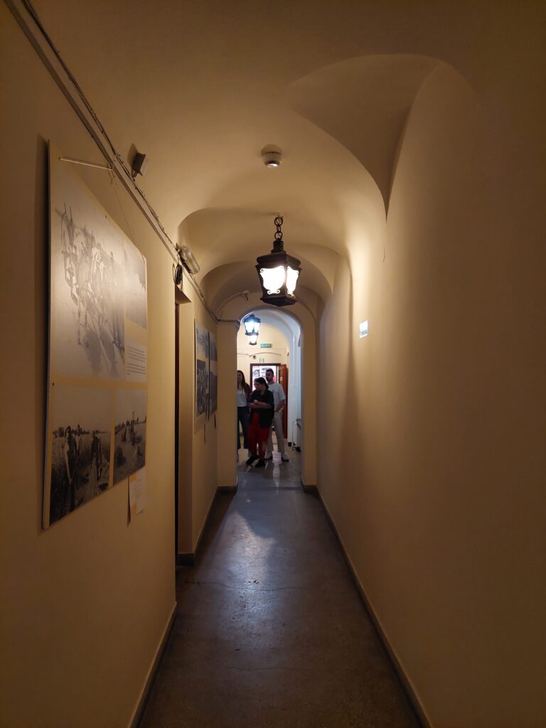 Wąski korytarz oświetlony lampą. Na ścianie wisząca plansza.