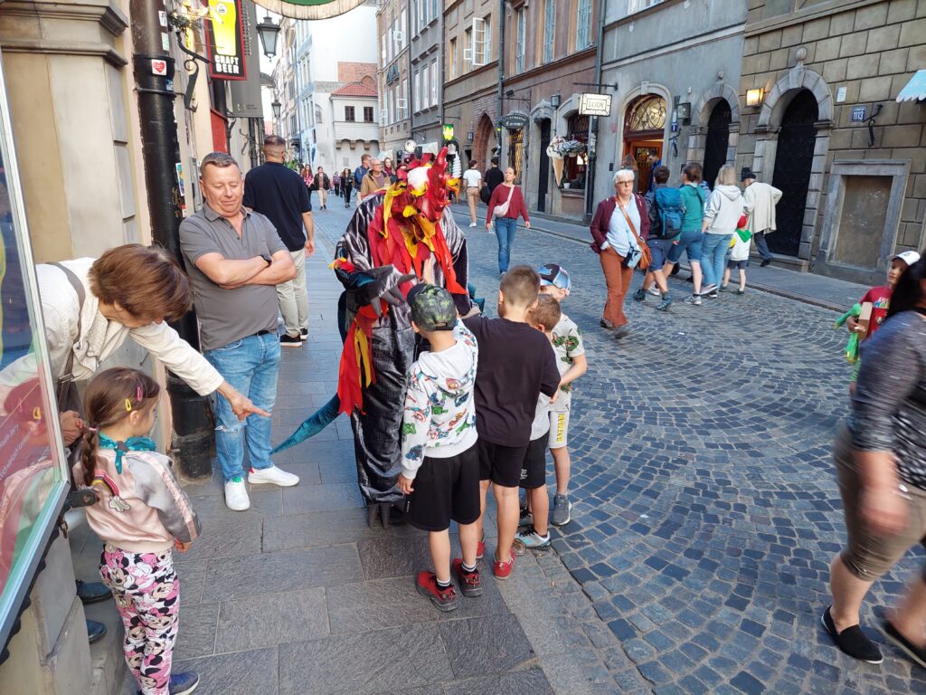 Grupa dzieci i dorosłych z Bazyliszkiem na ulicy Nowomiejskiej na tle kamienic.