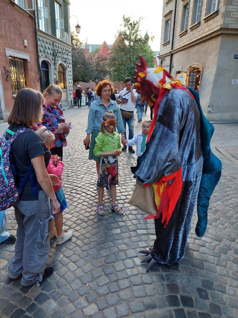 Grupa dzieci i dorosłych z Bazyliszkiem na ulicy Nowomiejskiej na tle Barbakanu.