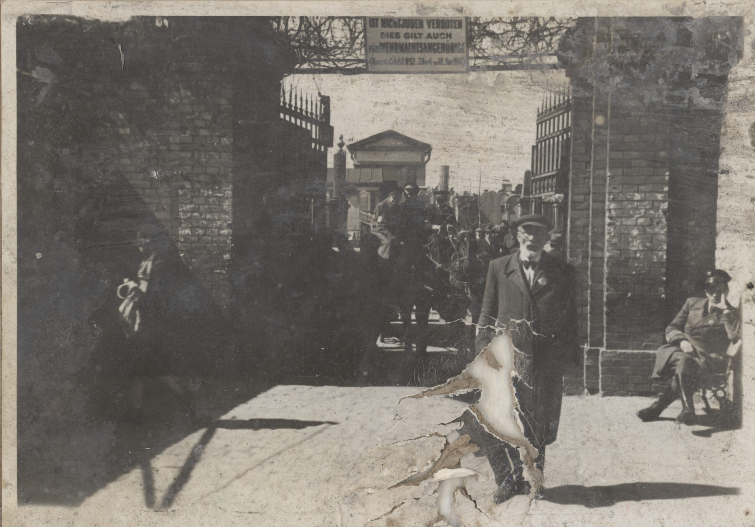Fotografia czarno-biała mężczyzny z brodą w kapeluszu i płaszczu, stojącego przed bramą Cmentarza Żydowskiego. Za nim widoczni funkcjonariuszy Żydowskiej Służby Porządkowej.
