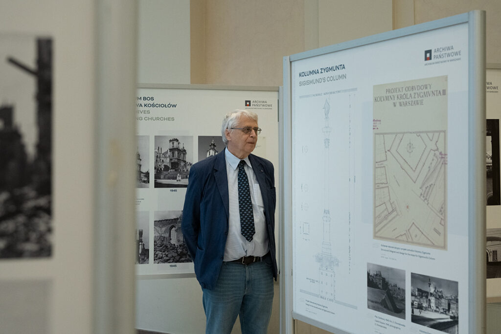 Fotografia starszego mężczyzny w krawacie stojącego między planszami wystawowymi