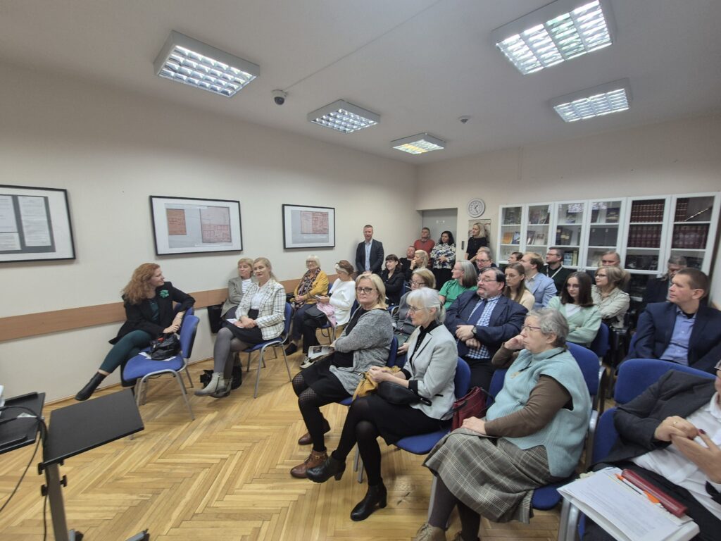 Fotografia grupy ludzi siedzących na krzesłach wewnątrz sali. Na ścianach wiszą obrazy w antyramach. Zdjęcie z lewego profilu.