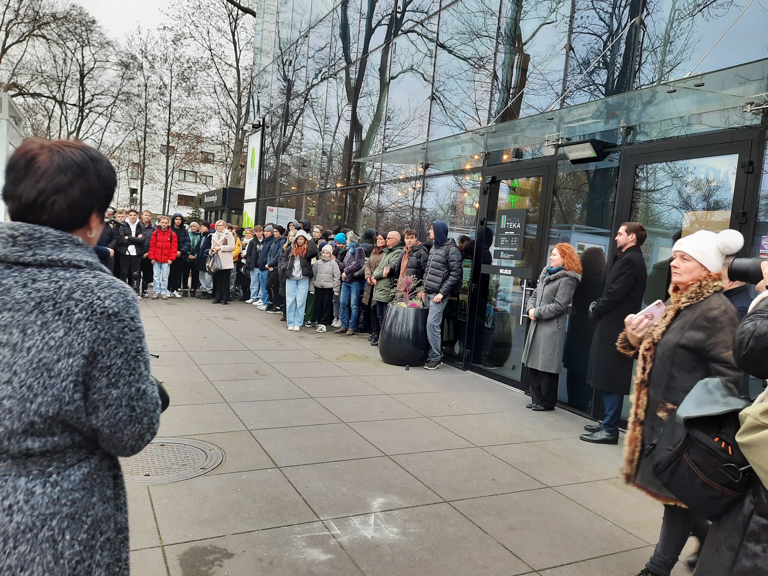 Fotografia kolorowa ludzi stojących między przeszklonym budynkiem a planszami wystawowymi w kubikach.