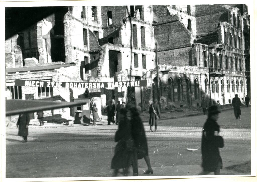 Na czarno-białej fotografii widać ruiny budowli, transparent z napisem „Niech żyje bohaterska Warszawa” i spacerujących ludzi.