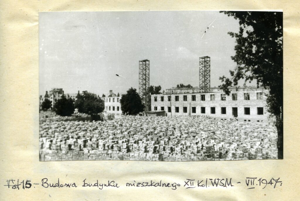 Na czarno-białej fotografii widać wiele cegieł i odbudowywany budynek w tle z dwoma masztami.