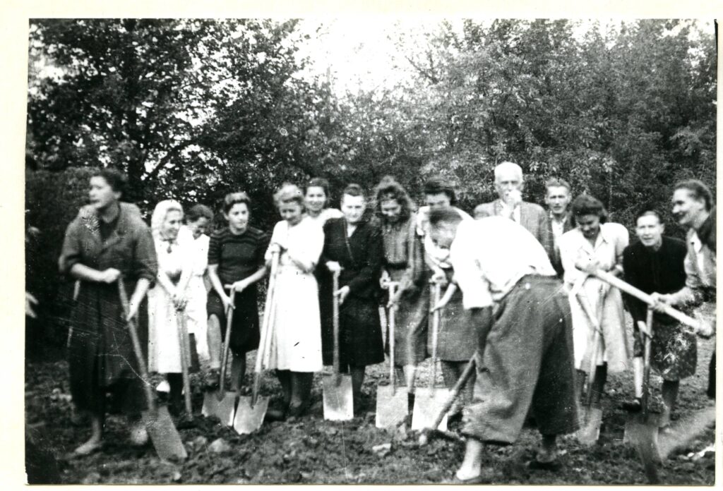 Na czarno-białej fotografii widać piętnaście osób z łopatami, na tle drzewa.