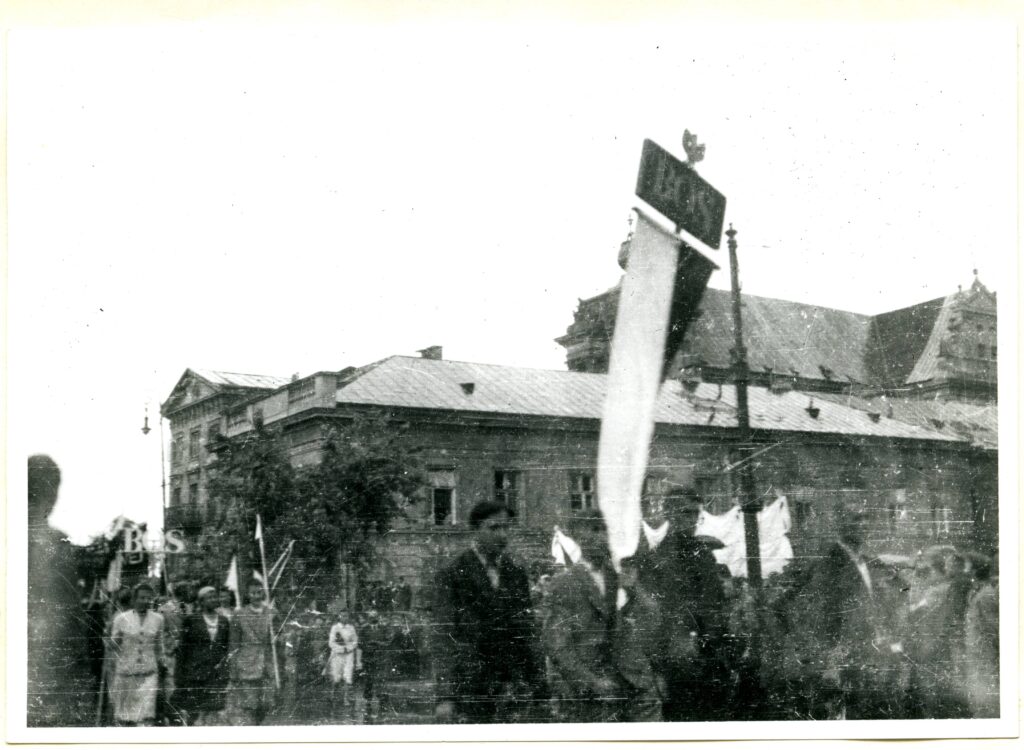 Na czarno-białej fotografii widać maszerujących ludzi ze sztandarem z napisem „BOS”, w tle również widać napis „BOS”.