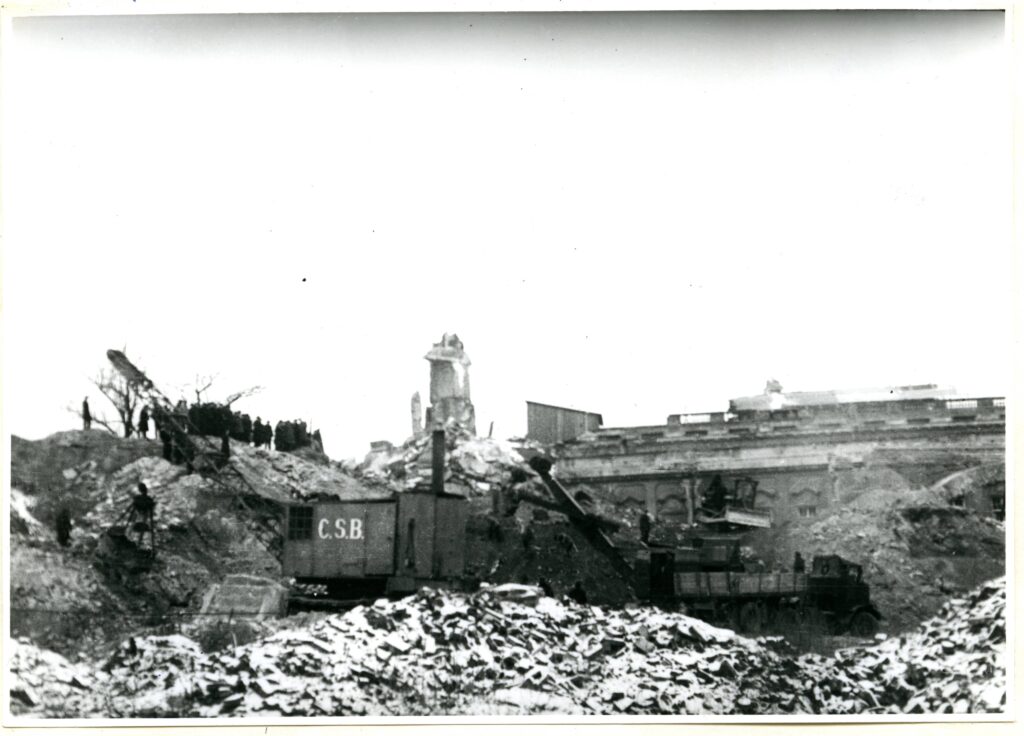 Na czarno-białej fotografii widać grupę ludzi, pojazdy (jeden ze oznakowaniem C.S.B.) i sprzęt budowlany wśród ruin.