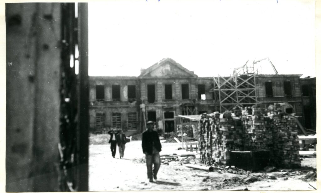 Na czarno-białej fotografii widać trzech mężczyzn, cegły i zrujnowany budynek.