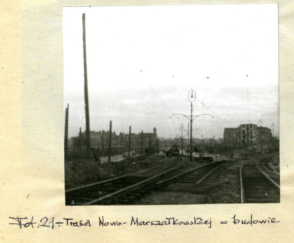 Na czarno-białej fotografii widać tory kolejowe z siecią trakcyjną, a w tle budynki.