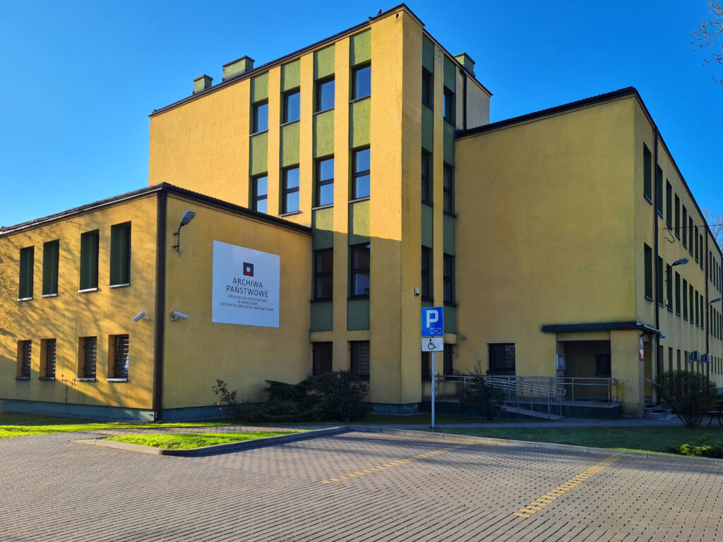 Kolorowa fotografia elewacji żółtego budynku z białą tablicą. Widoczny znak parkingu i kilka miejsc do parkowania.