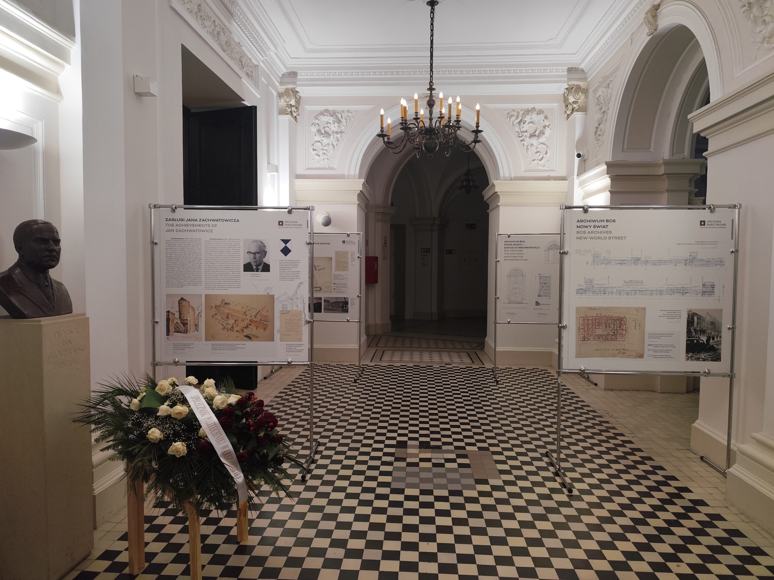 Na kolorowej fotografii widać plansze wystawiennicze, popiersie mężczyzny, wieniec i eleganckie wnętrze.