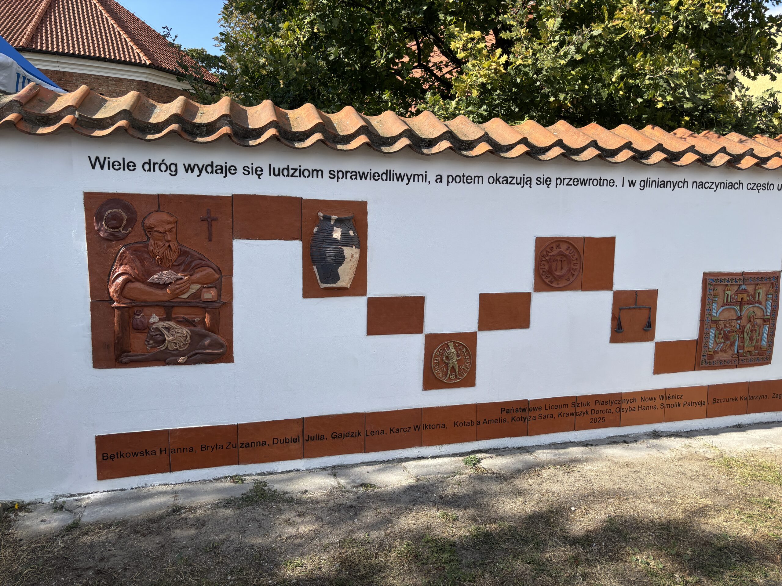 Na zdjęciu widać kompozycję ceramicznych płaskorzeźb, w tle drzewa i budynki