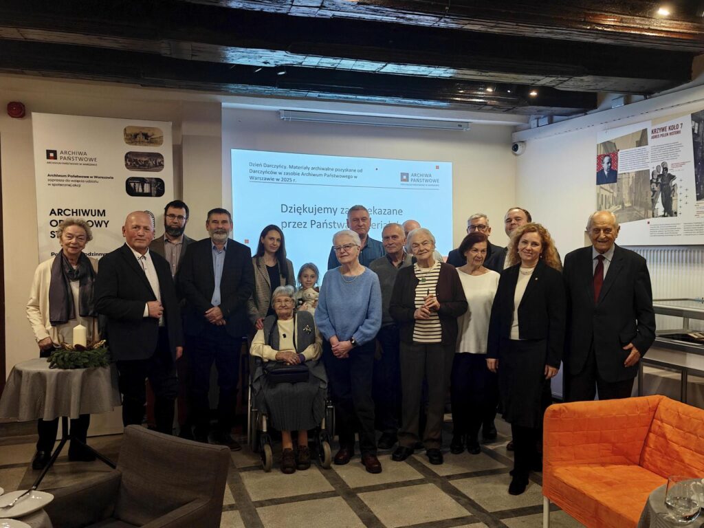 Na kolorowej fotografii widać siedemnaście osób w różnym wieku pozujących do zdjęcia na tle kasetonu i prezentacji, przed nimi widać fotele i gabloty.