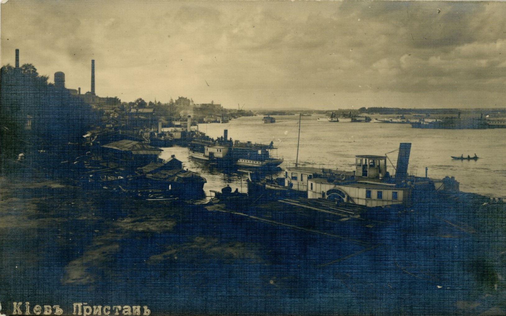 Pocztówka ukazująca przystań w Kijowie, 1917 r., Dar Macieja Aleksiejuka, Archiwum Państwowe w Warszawie, sygn. 72/3882/0/24/1603 Czarno-białe zdjęcie ukazujące brzeg z małymi domkami i przybijające do brzegu statki, w tle woda z pływającymi po niej statkami, w oddali także dwa kominy.
