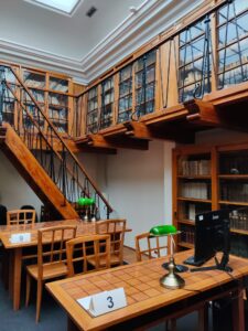 Na kolorowej fotografii widać dwa stoły, pięć krzeseł, komputer, schody i regały z książkami.