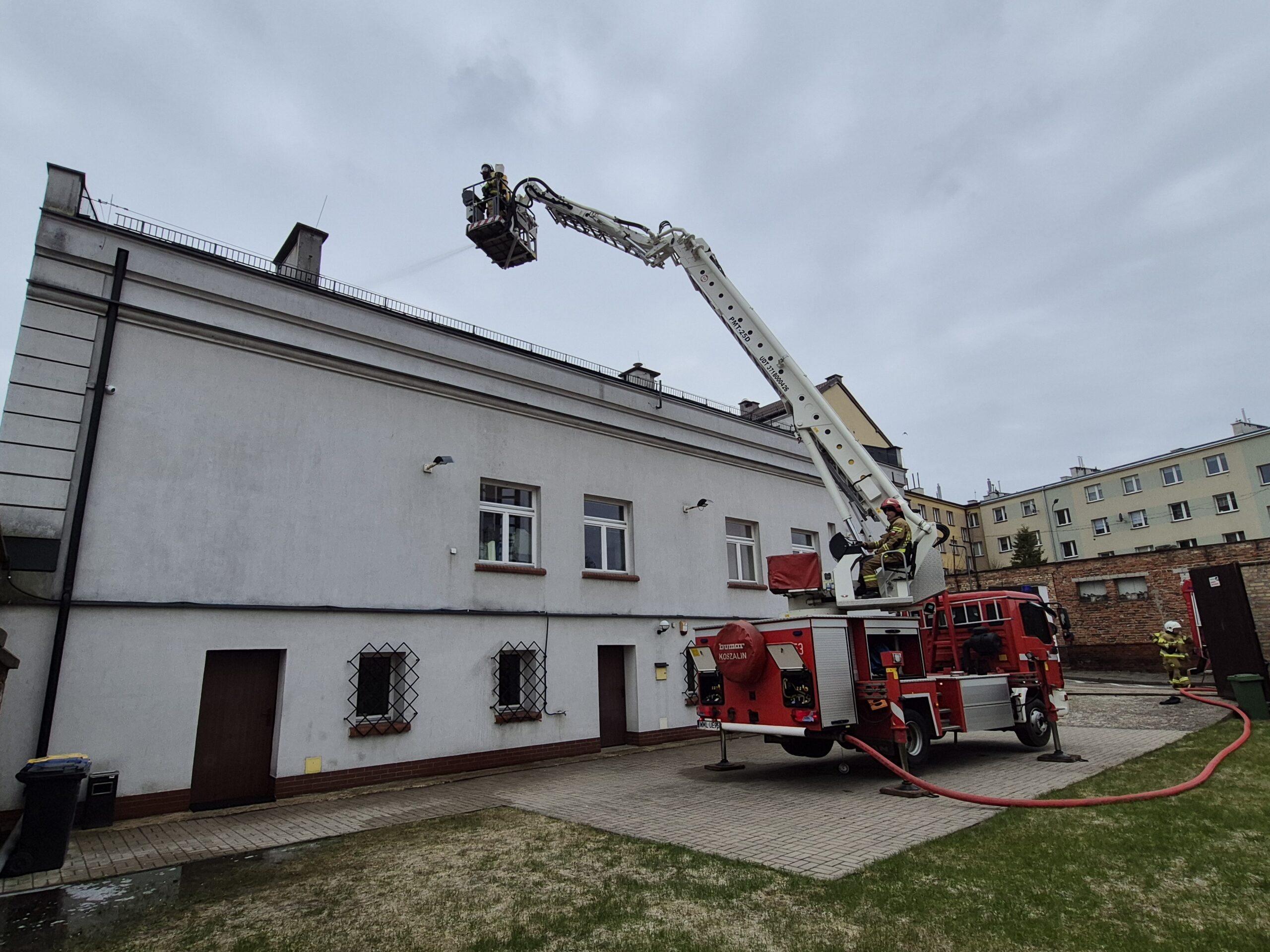 Na kolorowym zdjęciu widać trzech mężczyzn w strojach strażackich, z których jeden znajduje się na wysięgniku i gasi budynek, w tle widać mur i inny budynek.