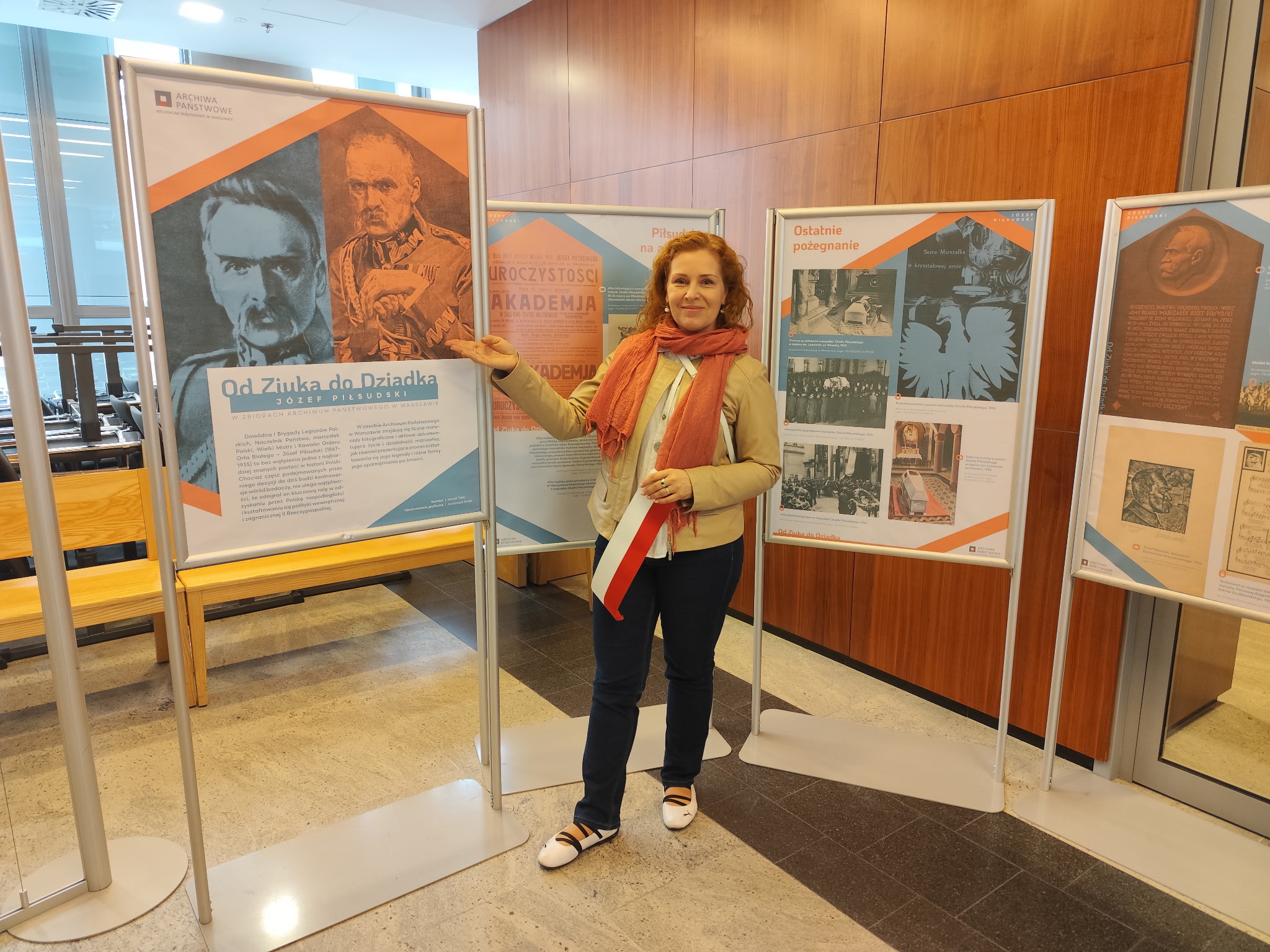 Dyrektor Archiwum Państwowego w Warszawie Magdalena Wiercińska z wystawą „Od Ziuka do Dziadka. Józef Piłsudski w zbiorach Archiwum Państwowego w Warszawie” na Nocy Muzeów 2026 w Naczelnym Sądzie Administracyjnym
Fot. M. Teler Na kolorowym zdjęciu widać rudowłosą kobietę trzymającą w ręku biało-czerwoną wstęgę na tle czterech tablic PCV.