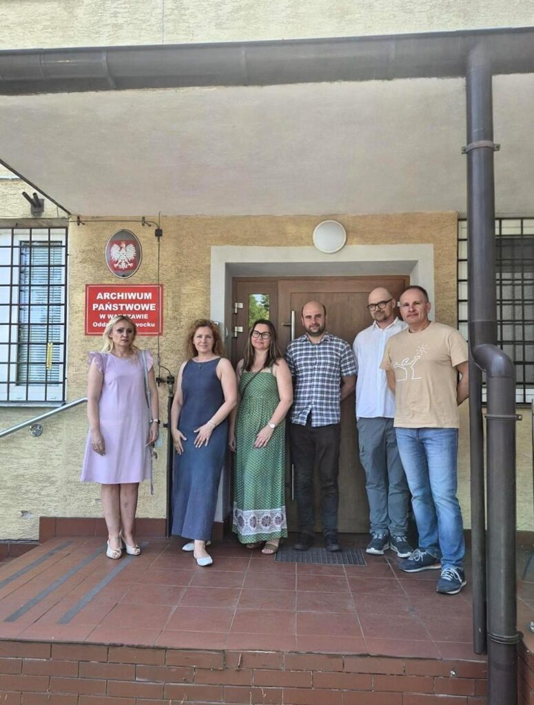 Na kolorowej fotografii widać trzy kobiety i trzech mężczyzn stojących przed budynkiem z godłem Orła Białego i czerwoną tablicą z białym napisem Archiwum Państwowe w Warszawie.