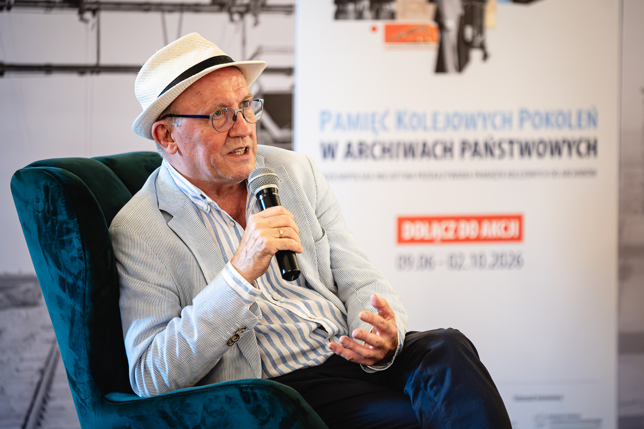 Aktor Artur Barciś na spotkaniu promującym nową akcję Archiwów Państwowych i PKP S.A. „Pamięć Kolejowych Pokoleń w Archiwach Państwowych”. Kolorowa fotografia ukazująca starszego mężczyznę w kapeluszu, w koszuli i marynarce, siedzącego na fotelu, w tle baner.