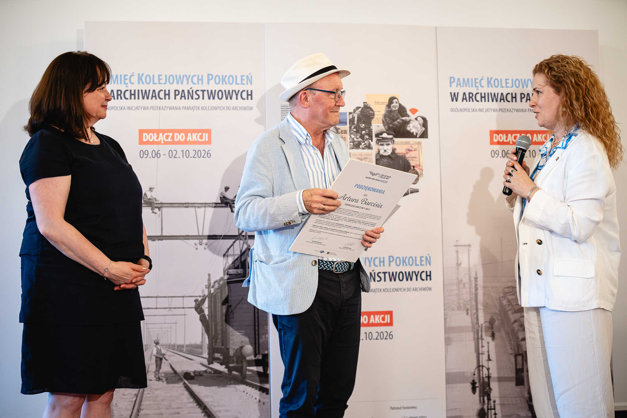 Artur Barciś odbiera podziękowania dla pierwszego darczyńcy akcji „Pamięć Kolejowych Pokoleń w Archiwach Państwowych” z rąk zastępcy Naczelnego Dyrektora Archiwów Państwowych dr hab. Lucyny Harc oraz dyrektora Archiwum Państwowego w Warszawie Magdaleny Wiercińskiej. Kolorowa fotografia ukazująca starszego mężczyznę w kapeluszu, w koszuli i marynarce, który odbiera dyplom od dwóch kobiet, jednej w czarnym kostiumie, drugiej w białej marynarce i koszuli w niebieskie paski, w tle ścianka.