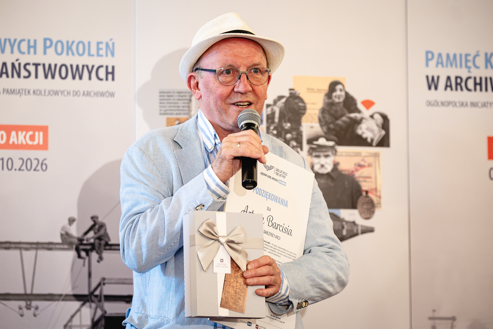 Aktor Artur Barciś na spotkaniu promującym nową akcję Archiwów Państwowych i PKP S.A. „Pamięć Kolejowych Pokoleń w Archiwach Państwowych”. Kolorowa fotografia ukazująca starszego mężczyznę w kapeluszu, trzymającego w ręku mikrofon oraz dyplom i upominek, w tle ścianka.
