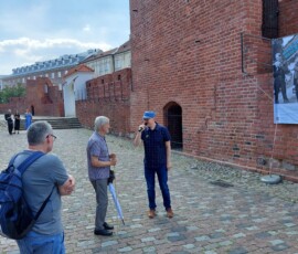 Fotografia trzech mężczyzn pod murem Barbakanu. Jeden w czapce z daszkiem trzyma mikrofon.