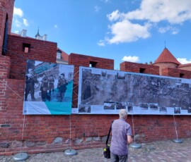 Fotografia mężczyzny oglądającego wystawę na barykadach. W tle widoczne ceglane mury Barbakanu i plansze wystawowe z tkanin.