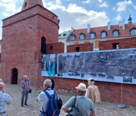 Fotografia grupy ludzi słuchającego starszego mężczyzny z mikrofonem pod murami Barbakanu z wiszącą wystawa z tkanin.