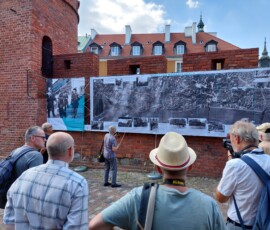 Fotografia tłumu ludzi słuchającego starszego mężczyzny z mikrofonem pod murami Barbakanu z wiszącą wystawa z tkanin.