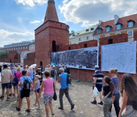 Fotografia tłumu ludzi słuchającego starszego mężczyzny z mikrofonem pod murami Barbakanu z wiszącą wystawa z tkanin.