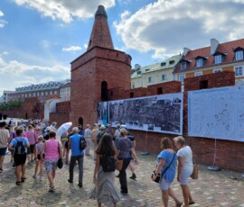 Fotografia tłumu ludzi słuchającego starszego mężczyzny z mikrofonem pod murami Barbakanu z wiszącą wystawa z tkanin.