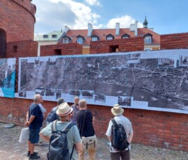 Fotografia tłumu ludzi słuchającego starszego mężczyzny z mikrofonem pod murami Barbakanu z wiszącą wystawa z tkanin.