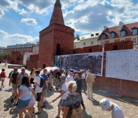 Fotografia tłumu ludzi słuchającego starszego mężczyzny z mikrofonem pod murami Barbakanu z wiszącą wystawa z tkanin.