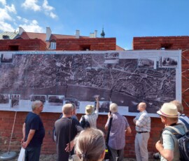 Fotografia tłumu ludzi słuchającego starszego mężczyzny z mikrofonem pod murami Barbakanu z wiszącą wystawa z tkanin.