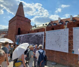 Fotografia tłumu ludzi słuchającego starszego mężczyzny z mikrofonem pod murami Barbakanu z wiszącą wystawa z tkanin. Widać dwa rozłożone, białe parasole.
