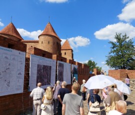 Fotografia tłumu ludzi słuchającego starszego mężczyzny z mikrofonem pod murami Barbakanu z wiszącą wystawa z tkanin. Widać dwa rozłożone, białe parasole.