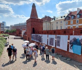 Fotografia tłumu ludzi słuchającego starszego mężczyzny z mikrofonem pod murami Barbakanu z wiszącą wystawa z tkanin. Widać trzy rozłożone, białe parasole. Zdjęcie zrobione z góry.