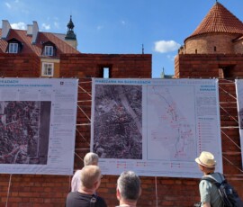Fotografia tłumu ludzi słuchającego starszego mężczyzny z mikrofonem pod murami Barbakanu z wiszącą wystawa z tkanin.