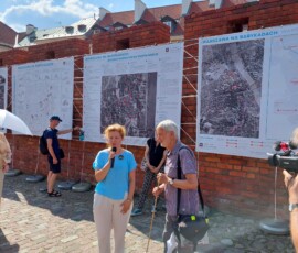 Fotografia kobiety z mikrofonem i starszego mężczyzny ze wskaźnikiem na tle murów Barbakanu i wystawy z tkanin.