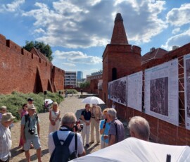 Fotografia tłumu ludzi słuchającego starszego mężczyzny z mikrofonem pod murami Barbakanu z wiszącą wystawa z tkanin.