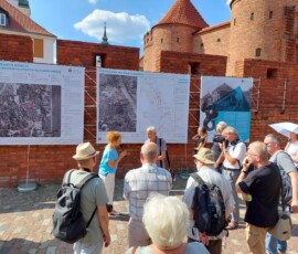 Fotografia kobiety z mikrofonem i starszego mężczyzny stojących w środku tłumu. Po prawej stronie zdjęcia mury Barbakanu i plansze wystawowe z tkanin. Po lewej stronie pas zieleni i mur.