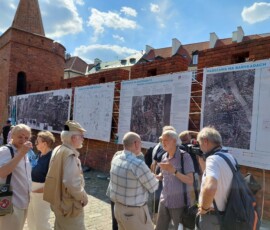 Fotografia starszego mężczyzny z mikrofonem i wskaźnikiem, pokazującego na planszę. Za nim 5 osób, w tym dwie w kapeluszach. Wszyscy są odwróceni plecami do fotografa.