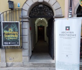 Kolorowa fotografia bramy wejściowej do kamienicy. Po prawej stronie roll up z logo APW, po lewej wiszący na kracie plakat na Urodziny Starówki.