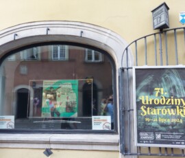 Kolorowa fotografia. Wiszący na kracie plakat na Urodziny Starówki, po lewej za szybą w witrynie telewizor z wyświetlającą się grafiką zapraszająca na wystawę "Warszawa na barykadach".