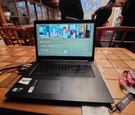 Fotografia laptopa z prezentacją multimedialną, leżącego na stole. W tle grupa ludzi.