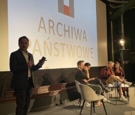 Dr Paweł Pietrzyk – Naczelny Dyrektor Archiwów Państwowych, Kamil Polak, Radosław Ładczuk, Hugh Welchman i Katarzyna Głuchowska – tłumaczka, fot. M. Frontczak Mężcyzna z mikrofonem na pierwszym planie. Na drugim planie siedzi trzech mężczyzn i kobieta. W tle ekran z logo Archiwów Państwowych z prostokątami.