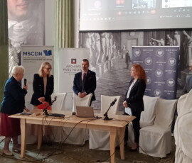 Fotografia 4 osób stojących za stołem prezydialnym między zielonymi kolumnami.