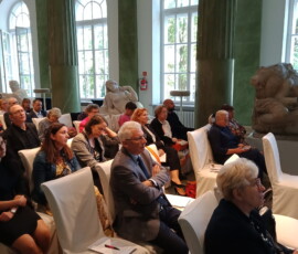 Fotografia grupy słuchaczy siedzących na białych krzesłach w sali z zielonymi kolumnami.
