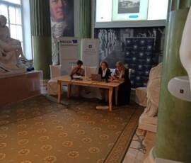 Fotografia 3 kobiet siedzących za stołem prezydialnym. Nad stołem widoczny telebim z prezentacją. Jedna kobieta trzyma mikrofon. Kadr z daleka.